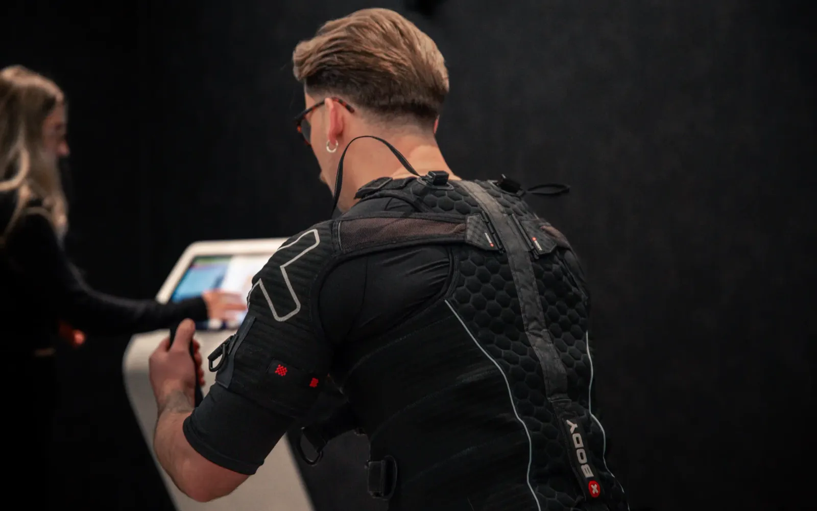 A man viewed from behind wearing a black EMS training suit fitted with electrode pads