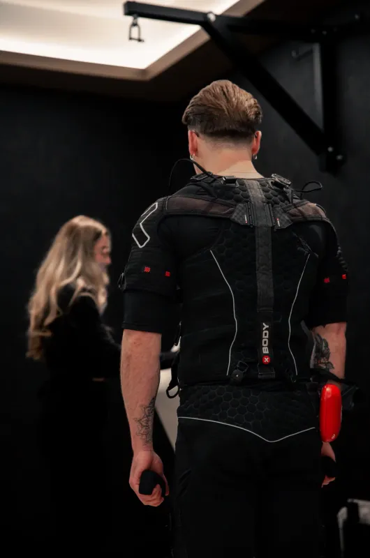 A man viewed from behind wearing a full EMS training suit with a red control unit at his waist, a trainer in the background