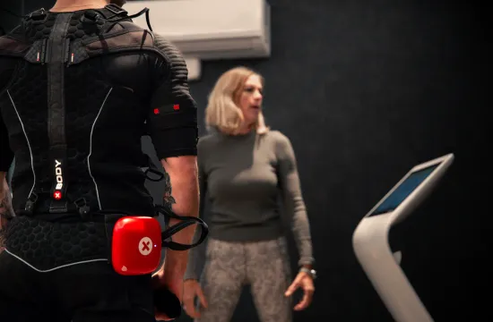 A man wearing an EMS suit with a red control unit at his waist, talking with a trainer near a treadmill