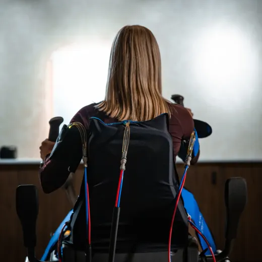 A woman seated on the Vasper System is seen from behind, wearing a black compression vest connected by cables to the machine