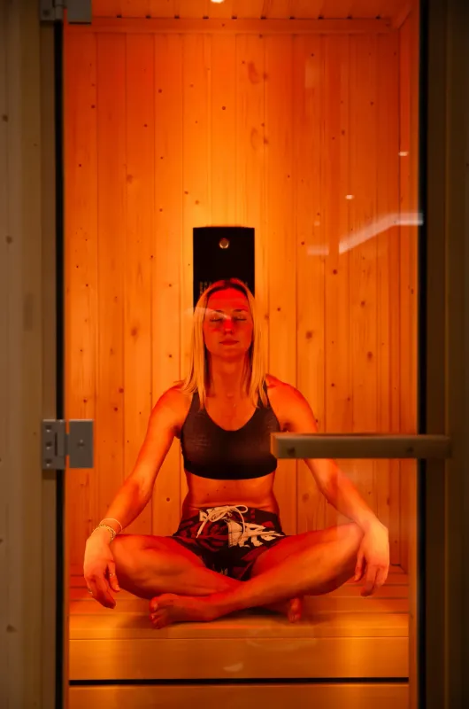 A woman sits cross-legged with her eyes closed in the infrared sauna, viewed through the open glass door