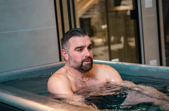 A man sitting in a cold plunge tub, looking to the side, in a modern studio setting