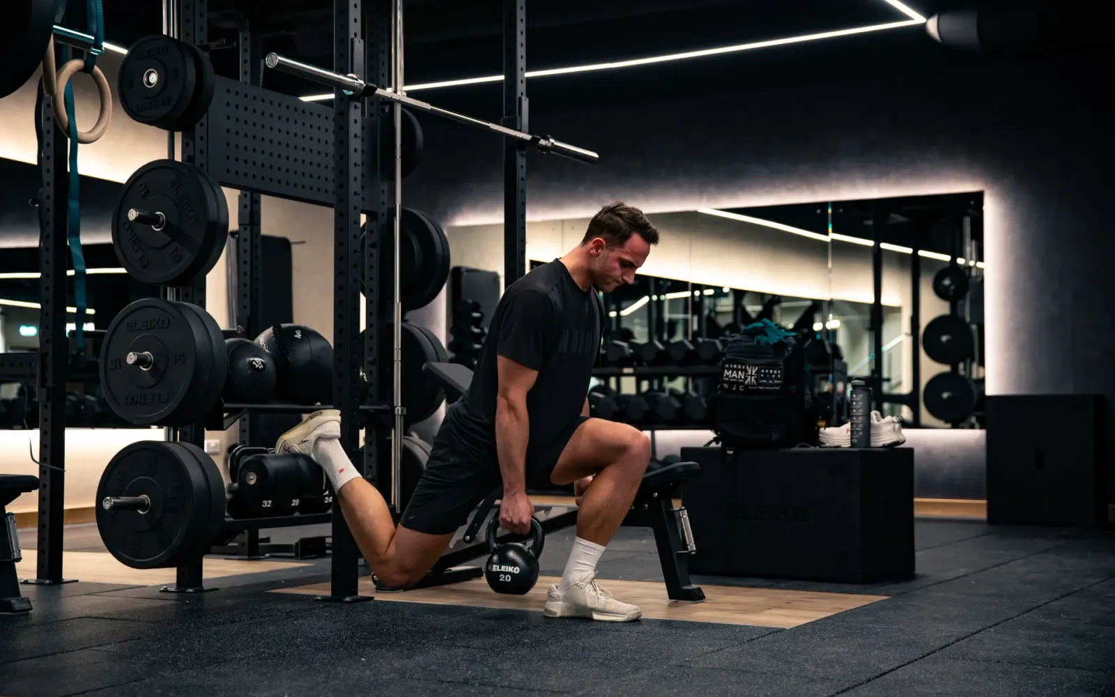 A man performing a single-leg kettlebell exercise on a bench in a dark, well-equipped gym
