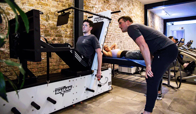 Two trainers assist a client using a Balkan Fitness strength training machine in a brick-walled gym