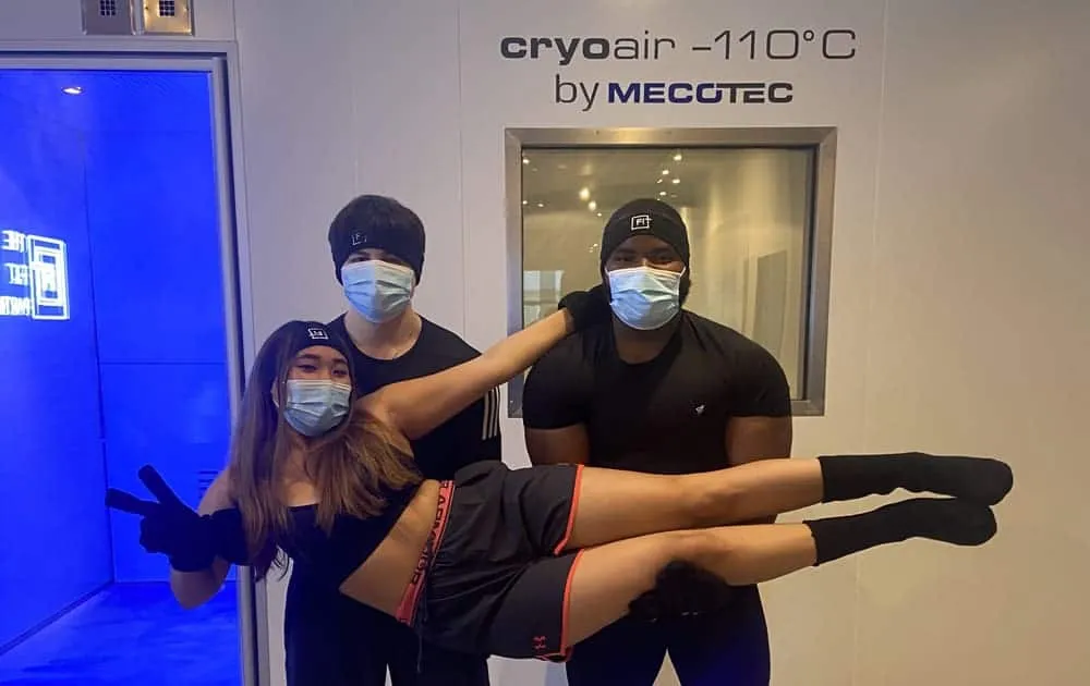 Three people wearing face masks and beanies pose beside a cryotherapy chamber marked cryoair -110C by Mecotec