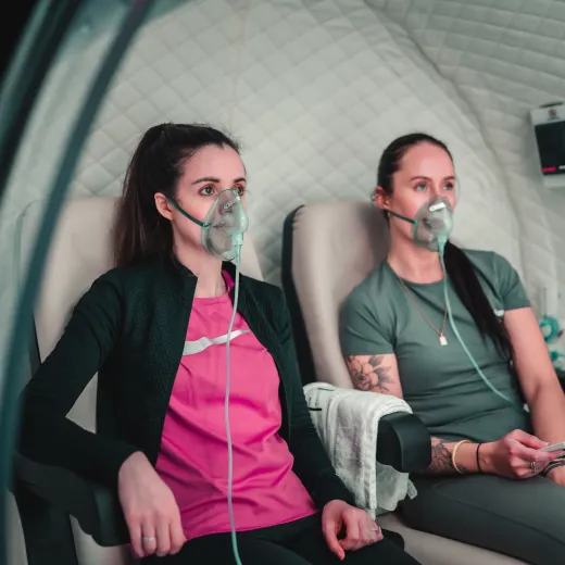 Two women sit side by side wearing oxygen masks inside a hyperbaric chamber, with a monitoring control panel visible behind them
