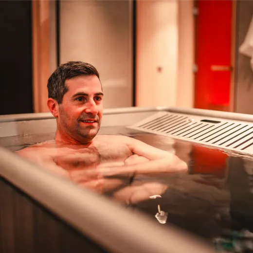 A man sitting in a cold plunge tub with a towel and red sauna door visible behind him