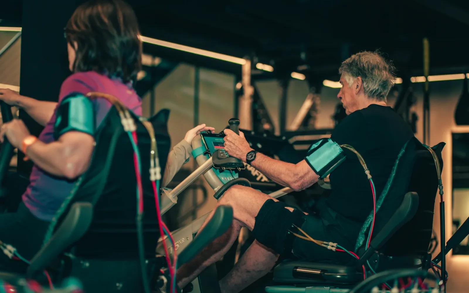 Two men seated on Vasper System machines with compression cuffs on their arms, gripping the handles during a session at The FIT Partnership