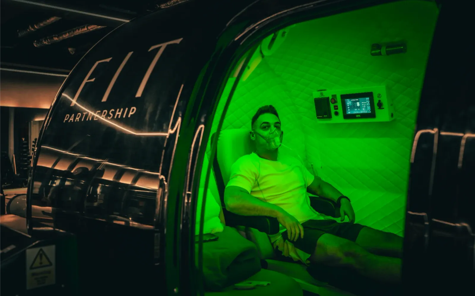A client reclines inside a hyperbaric oxygen chamber branded with the FIT Partnership logo, wearing an oxygen mask under green interior lighting
