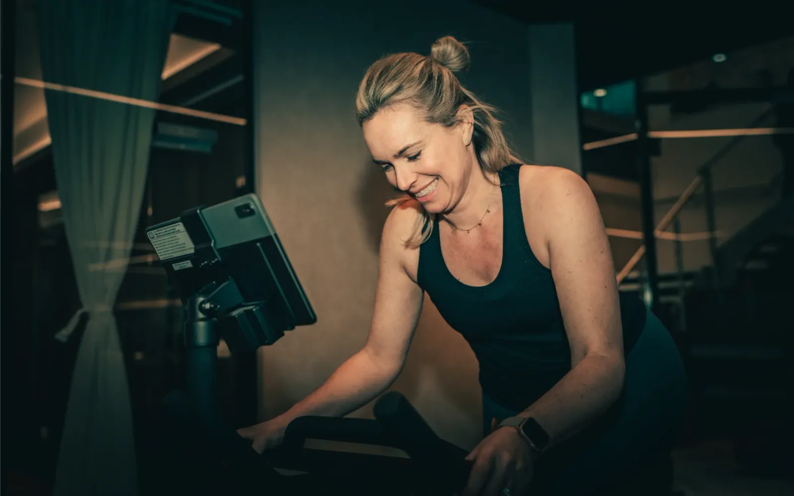 A smiling woman cycling on a CAROL bike in a dimly lit gym, looking at the console screen