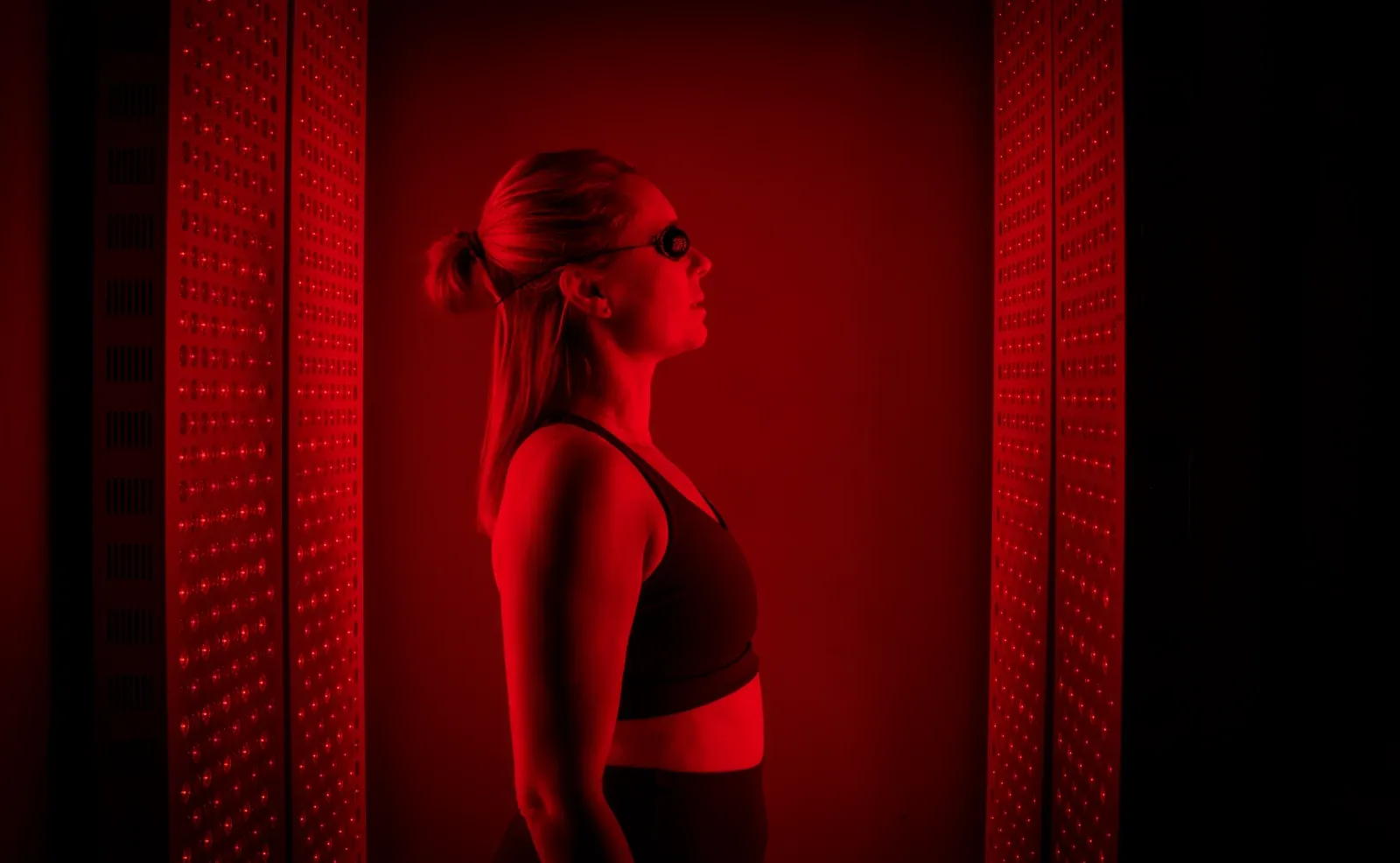 A woman wearing protective goggles stands in profile between two red light therapy panels glowing red