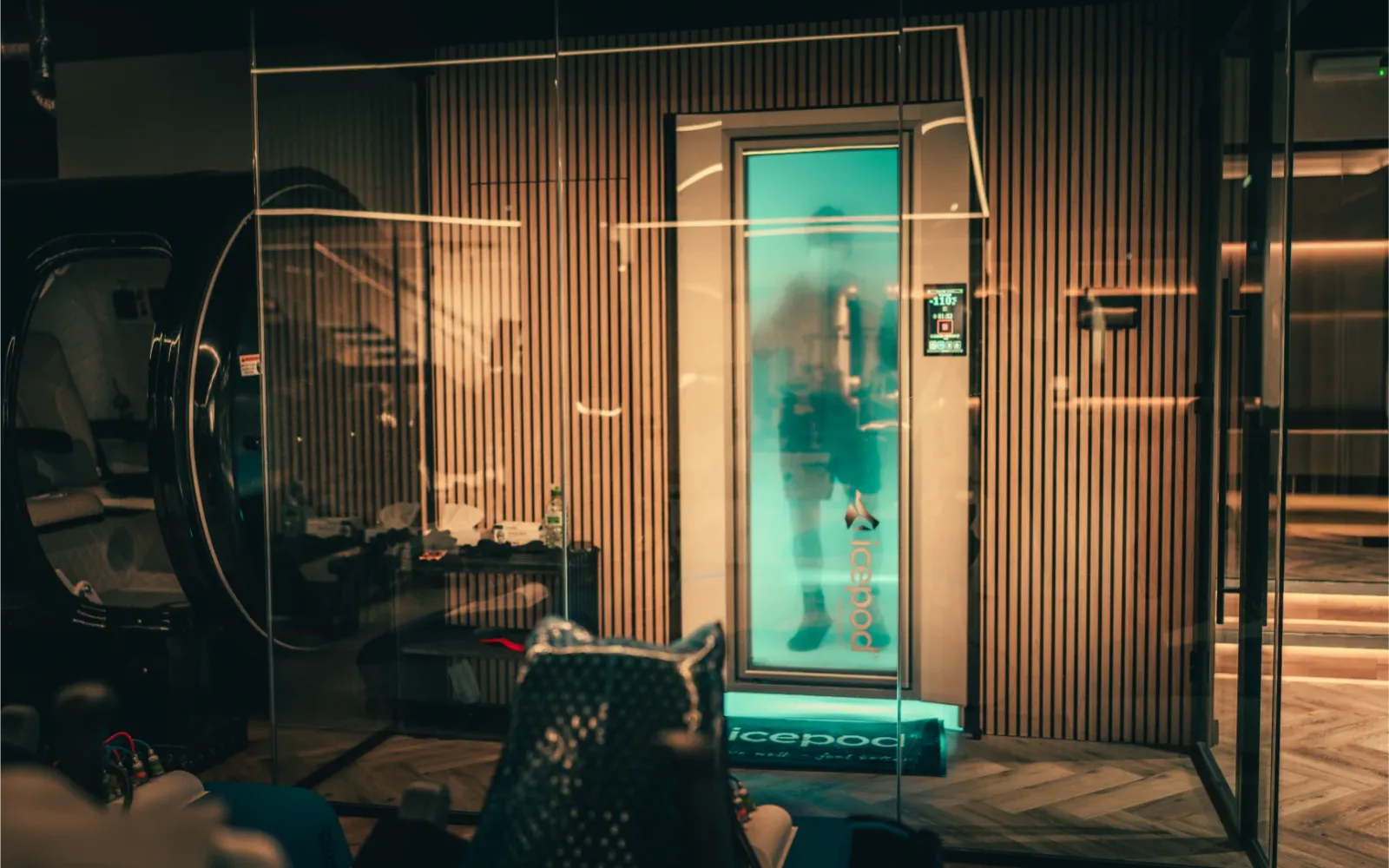 Exterior of the icepod cryotherapy chamber with a person visible inside through the glowing teal door