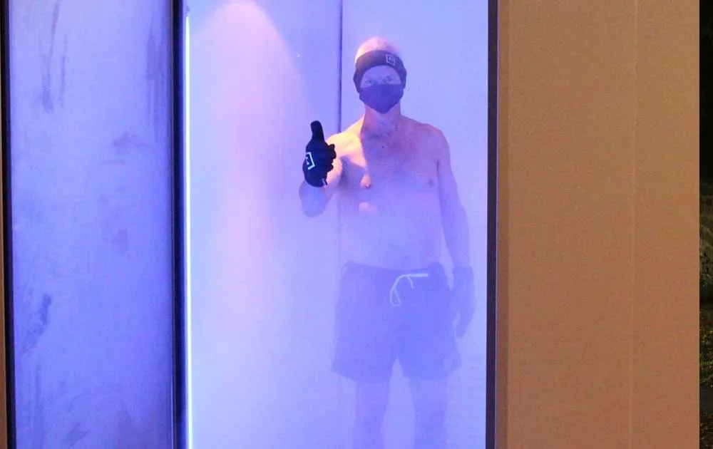 A man gives a thumbs up while standing inside a misty blue-lit cryotherapy chamber