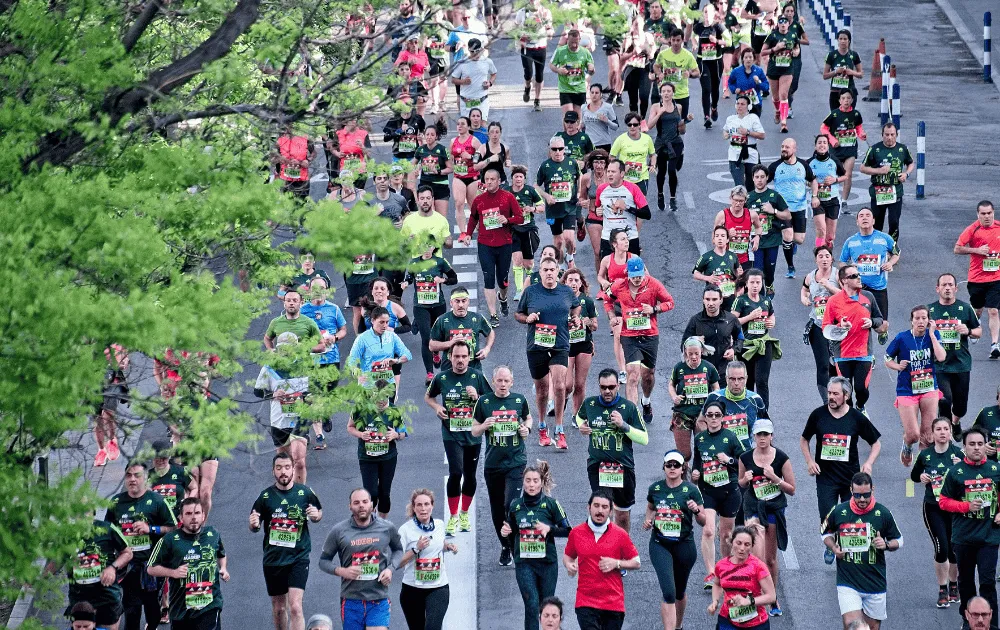 A dense crowd of marathon runners in branded race shirts running along a tree-lined street