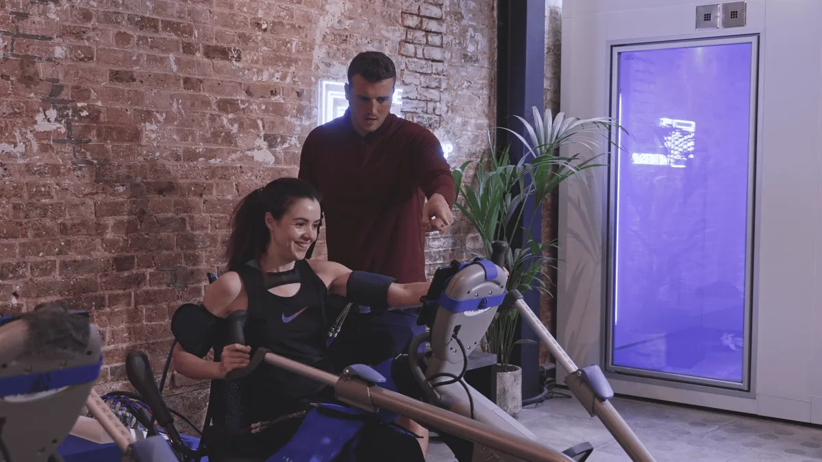 A trainer guides a woman using a Vasper exercise machine in a brick-walled studio