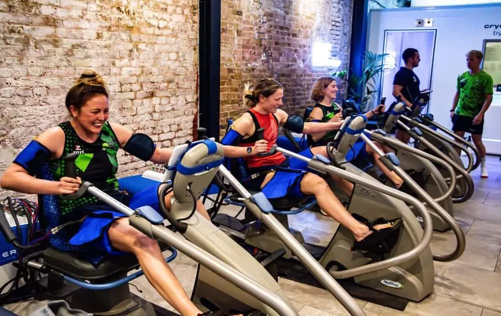 Group of people using Vasper compression exercise machines lined up against a brick wall
