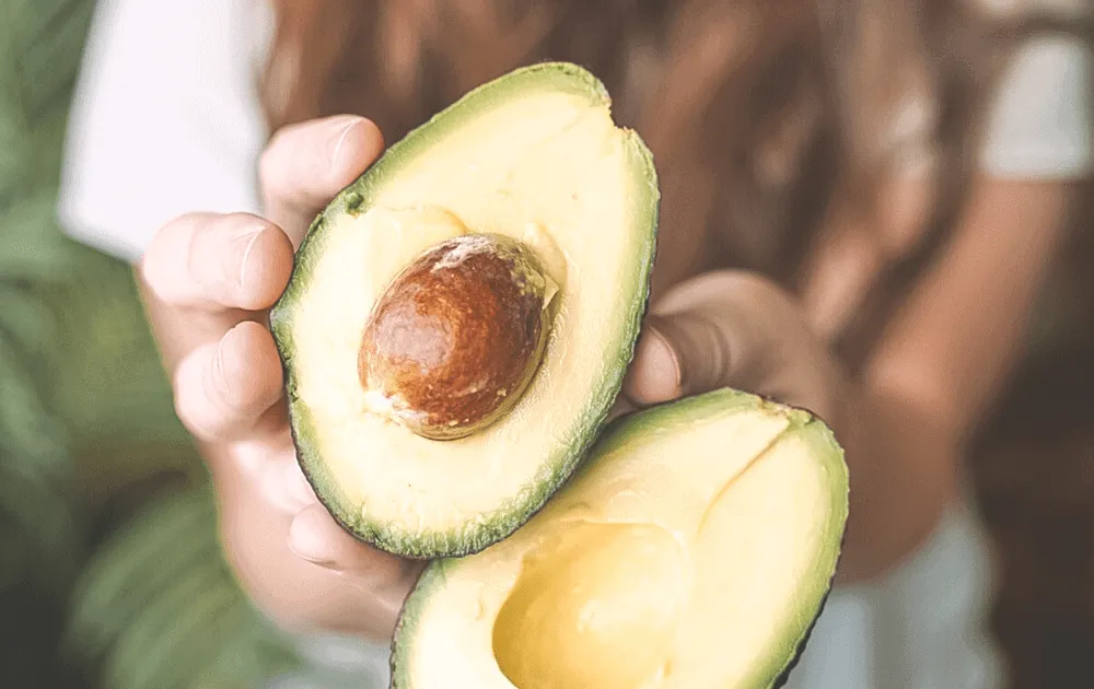 A person holds two halves of a sliced avocado, one still containing the stone