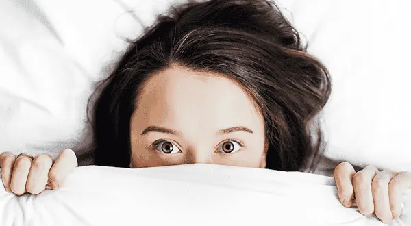 A woman peeks over a white duvet with only her eyes visible