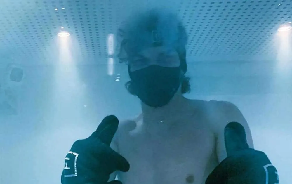A man gives a thumbs up inside a cryotherapy chamber wearing a face mask and gloves