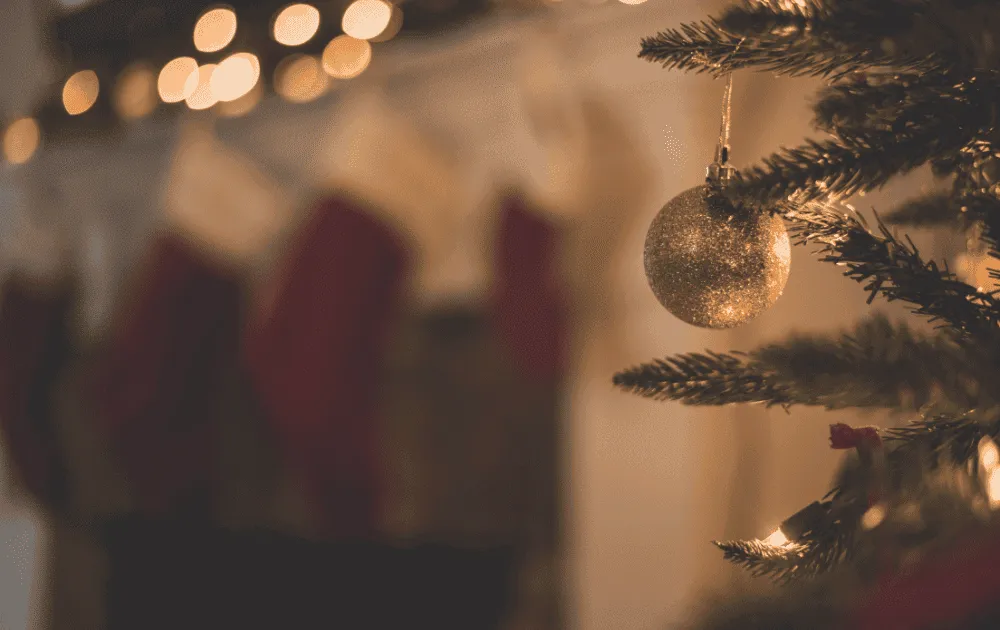 A glittering bauble hangs on a Christmas tree with fairy lights and stockings blurred in the background