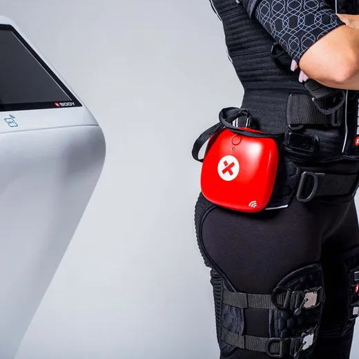 Close-up of a red EMS control unit clipped to the waistband of a training suit, beside a wall-mounted touchscreen