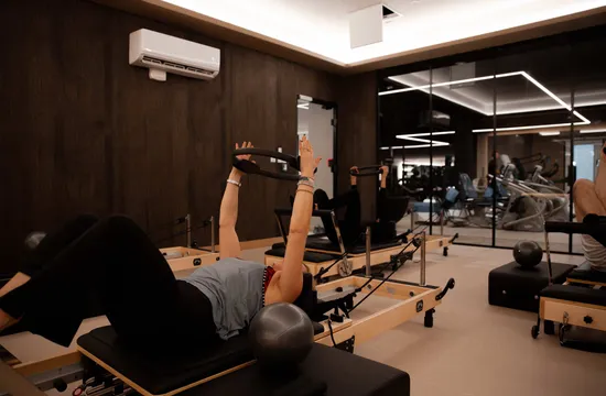 A client stretches on a Pilates reformer using an overhead ring, watched by other reformer users in the mirrored studio
