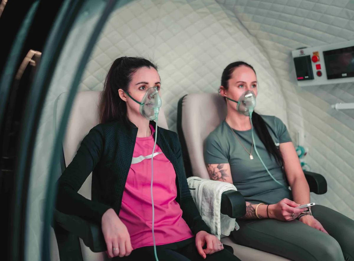 Two women sitting in a Hyperbaric Oxygen Chamber wearing oxygen masks