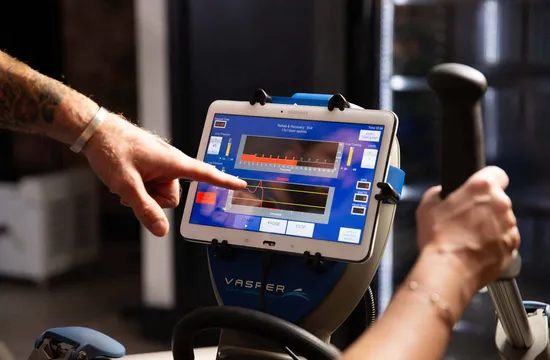 A tattooed hand points at the Vasper System's touchscreen display, showing interval timing, pressure and heart rate data mid-session