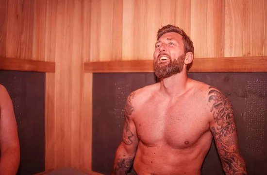 A bearded, tattooed man smiles and relaxes inside the wood-panelled infrared sauna at The FIT Partnership