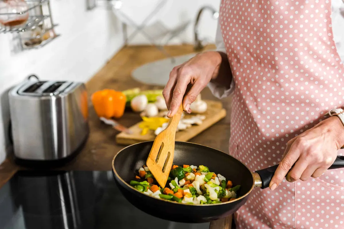 A hand uses a wooden spatula to stir chopped vegetables in a frying pan