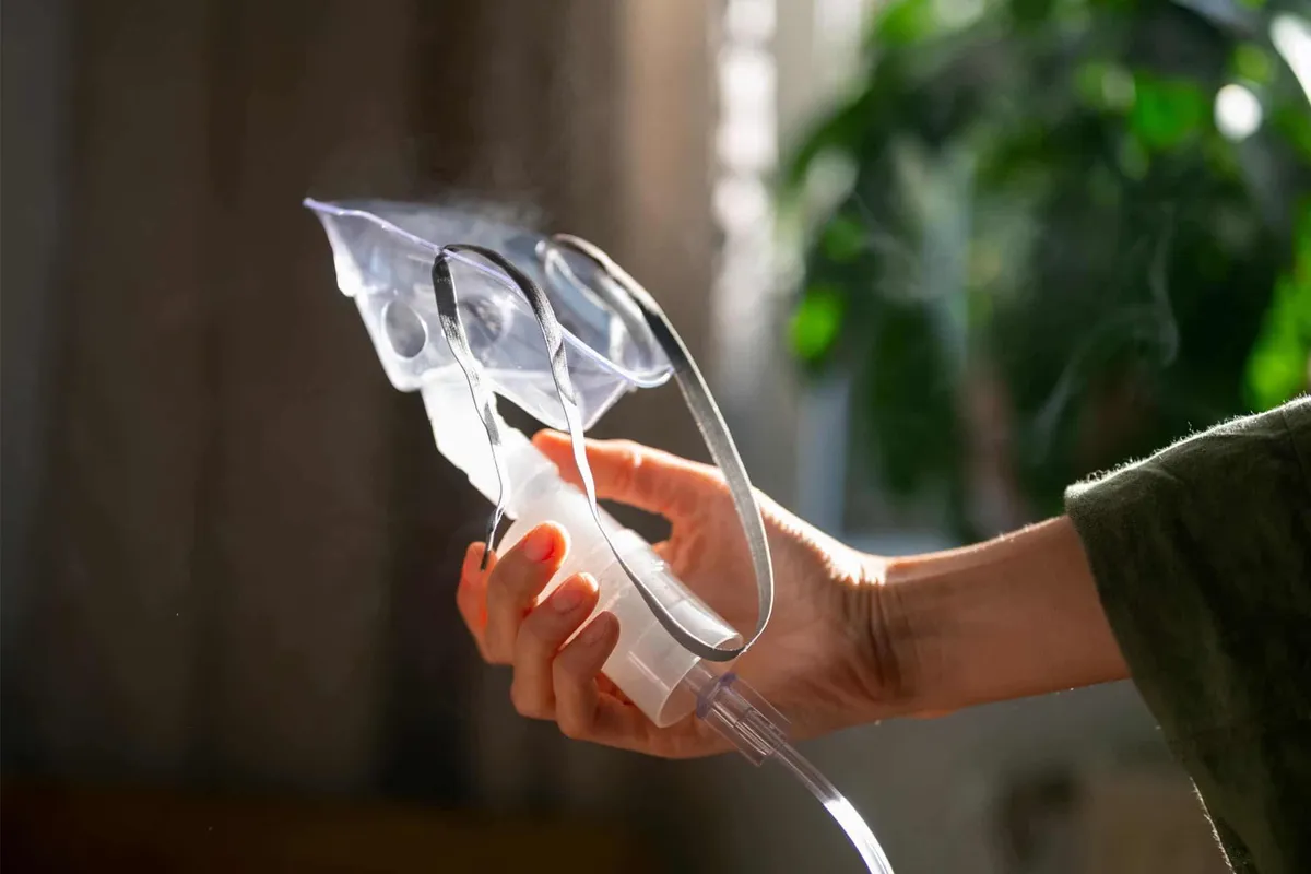 Hand holding a clear oxygen mask with attached tubing near a sunlit window