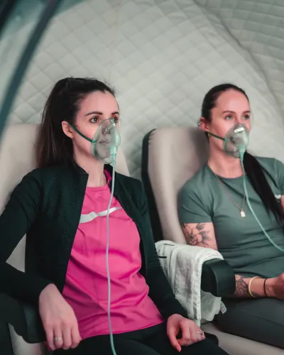 Two members wearing oxygen masks sit together during a duo hyperbaric oxygen therapy session