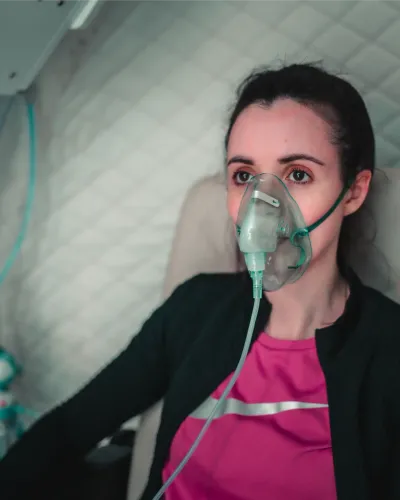 A woman wearing an oxygen mask sits inside a hyperbaric oxygen pod with its safety controls visible beside her
