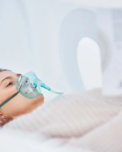 A woman reclines inside a hyperbaric oxygen pod wearing an oxygen mask, reaching toward the control panel