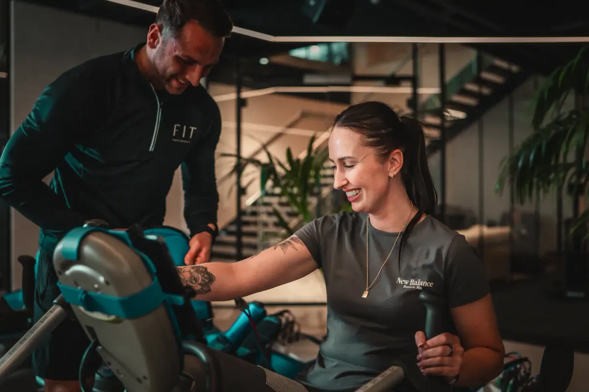 A FIT Partnership trainer guides a smiling member through a resistance machine exercise in the studio