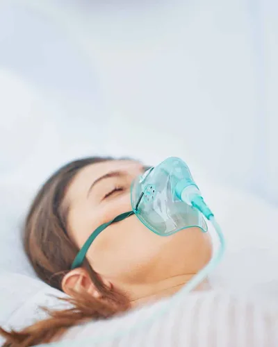 A woman lies back with eyes closed wearing an oxygen mask inside a hyperbaric therapy pod
