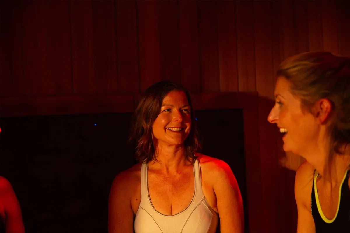 Two women smiling and chatting together inside a warmly lit infrared sauna