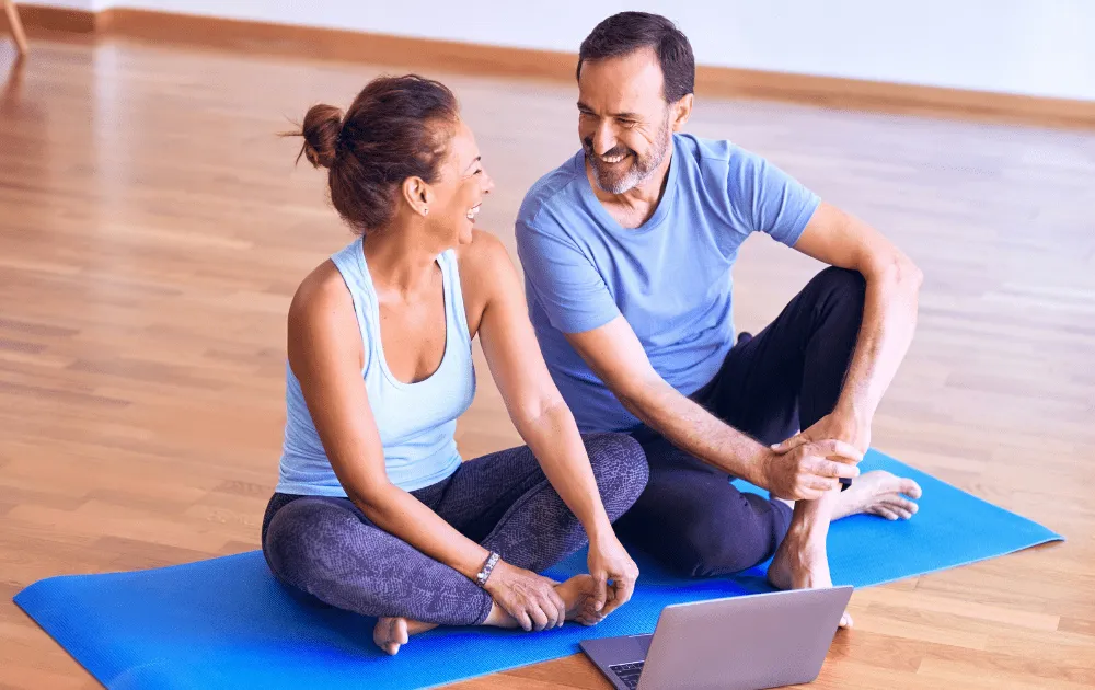 Smiling man and woman sitting cross-legged on a yoga mat chatting beside a laptop