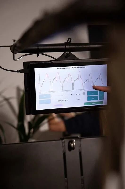 A finger points at the Balkan Strength Machine's touchscreen display, showing exercise repetition graphs during a session