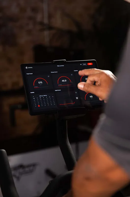 A hand tapping a CAROL bike console screen showing a ride summary with peak power and fitness score