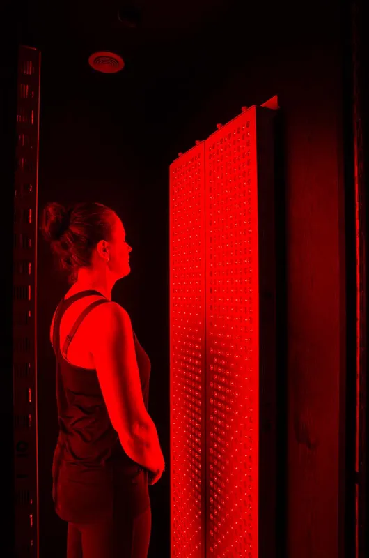 A woman stands side-on facing an illuminated red light therapy panel with her arms behind her back