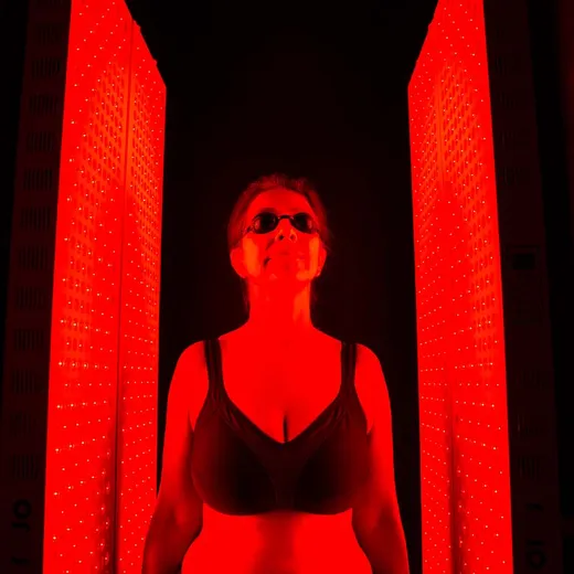 A woman with her hair in a bun and eyes closed relaxes in profile in front of a red light therapy panel