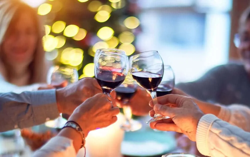 Hands clink wine glasses at a festive dinner table with a lit Christmas tree in the background