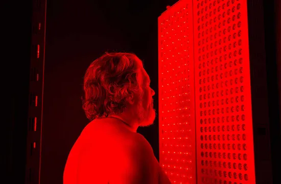 A bearded man stands shirtless in profile facing a glowing red light therapy panel