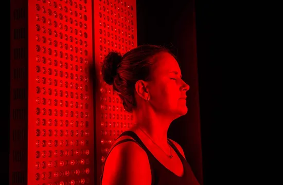 A woman wearing protective goggles stands facing forward between two illuminated red light therapy panels