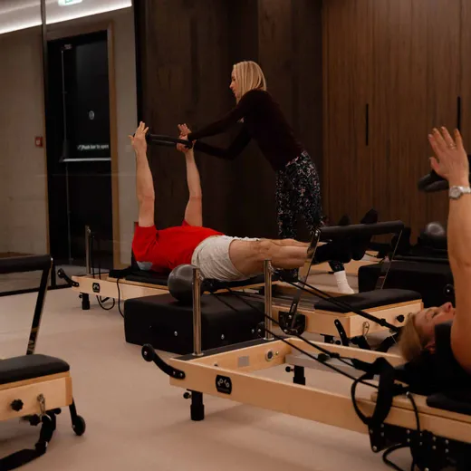 An instructor helps a client extend their arms overhead on a Pilates reformer using a resistance pole