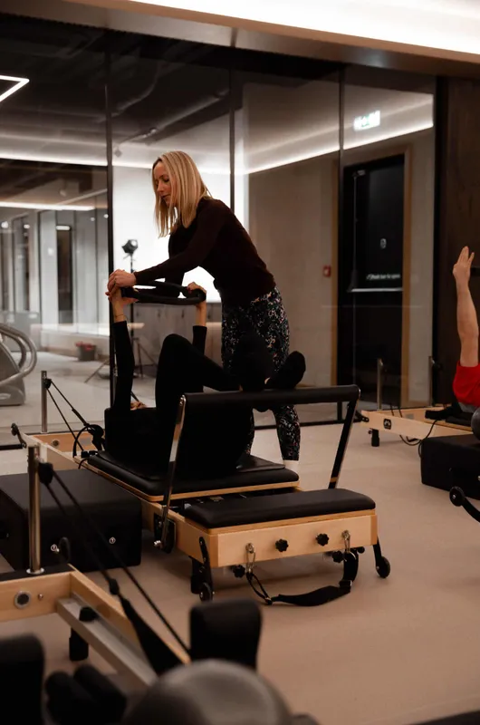 An instructor guides a client through a reformer exercise with bent knees, other members visible on nearby reformers