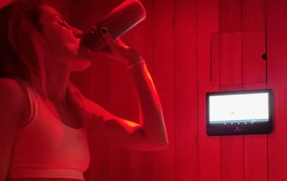 Woman drinking from a can inside a red-lit infrared sauna with a wall-mounted screen