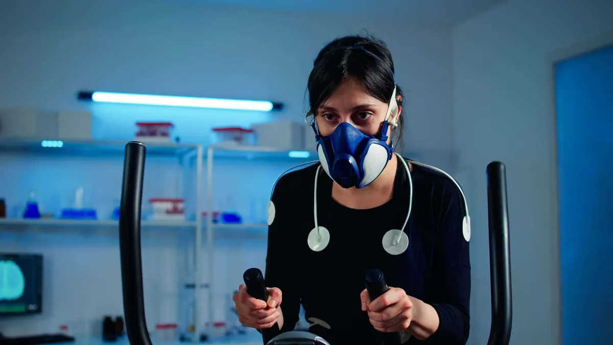 A woman wears a breathing mask and heart rate sensors while using an exercise bike in a lab setting