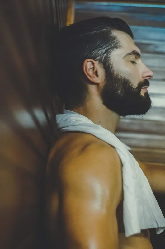 A man with his eyes closed and a towel around his neck relaxing on a sauna bench